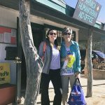 Two women wearing sunglasses on a sunny day smiling for a picture in front of a small grocery store.