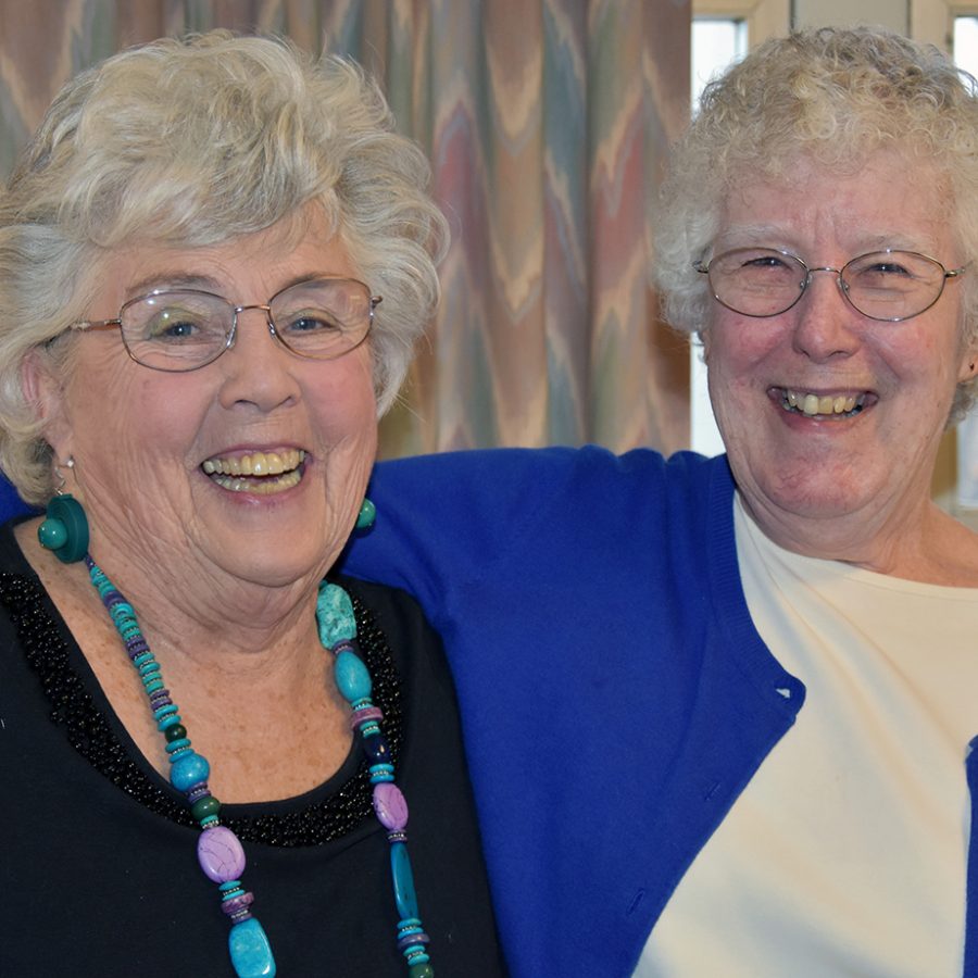 Two women, both with short grey hair and wire-framed glasses smiling brightly together for a picture together indoors. One woman has her arm around her friend's shoulder.