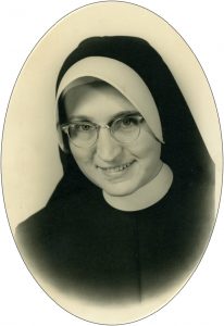 A black and white headshot of a nun wearing glasses and smiling.