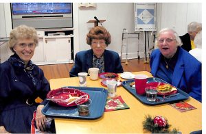 Three women smiling together at a table with lunch trays indoors.