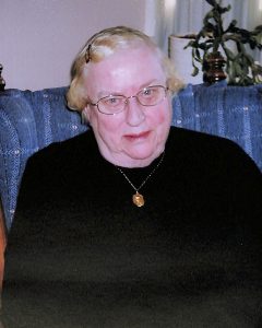 A woman with short blonde hair and glasses smiles for a picture in a black sweater while sitting on a blue plaid couch indoors.