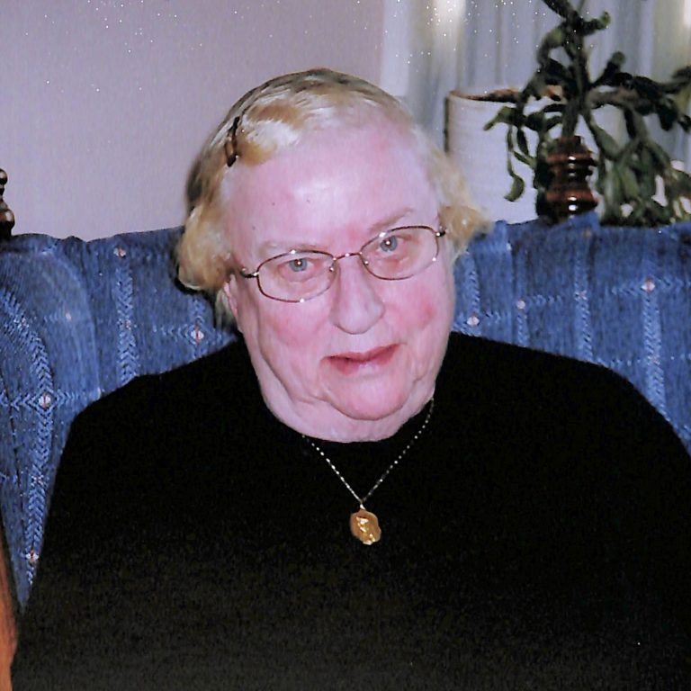 A woman with short blonde hair and glasses smiles for a picture in a black sweater while sitting on a blue plaid couch indoors.