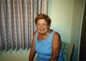 A woman with short brown hair and glasses wearing a blue tank top smiling for a picture while sitting on a chair indoors.