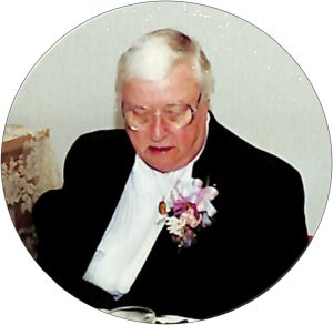 A woman with short grey hair and big glasses looking down while wearing a black blazer, white shirt, and flower boutonnière.
