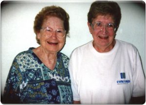 Two women smiling brightly together for a picture. Both women have short brown hair and glasses, one woman is wearing a blue patterned blouse, and one woman wearing a white t-shirt.