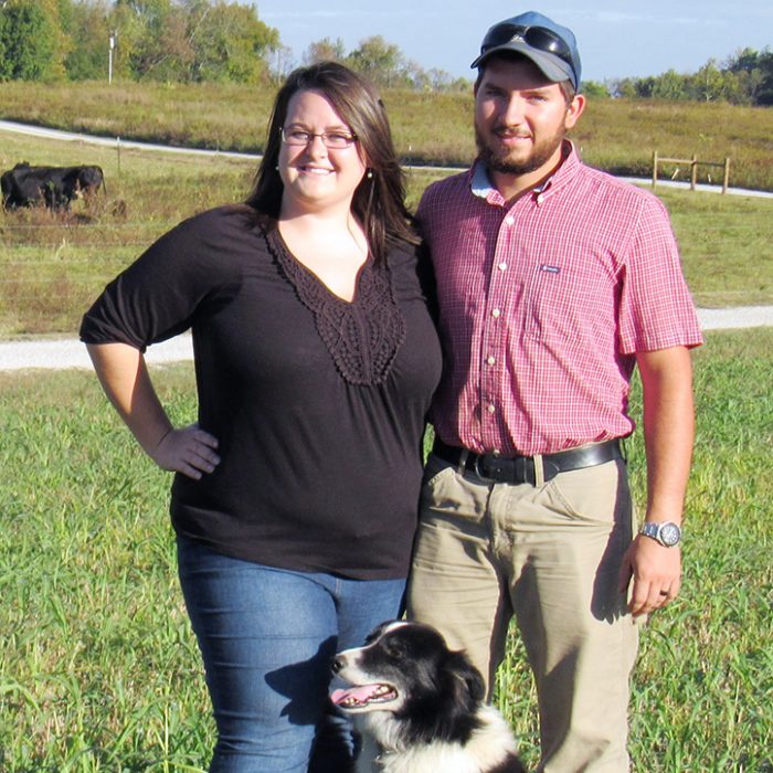 A man and woman smile for a picture together with a black and white border collie in a farm pasture on a sunny day.