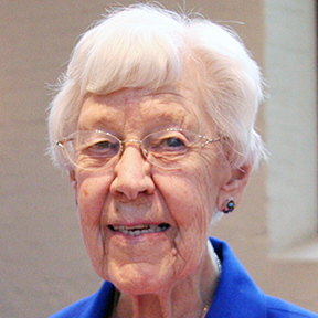 A woman with short, white hair, and round wire glasses, smiling for a headshot picture wearing earrings and a royal blue collared shirt.