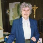 A woman standing in a religious classroom with a cross and a map in the back, the woman is smiling for a picture, with short grey hair, and big glasses, wearing a white blouse, a dark blue blazer.