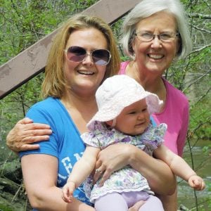 A grandmother, mother, and infant daughter smiling together for a group picture outdoors on a sunny day.