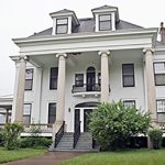 A grand white mansion with four columns in the front and lush green grass, bushes, and trees surrounding it.