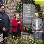 Seven individuals were pictured candidly in front of a plaque on a red brick building on an overcast day.
