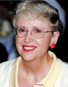 A woman with short, light colored hair smiling brightly for a picture, wearing a light yellow collared blouse, red lip stick, round wire glasses, and red lipstick.
