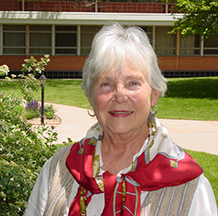 A woman with short, white hair wearing a white blouse and a red patterned scarf while smiling brightly outdoors on a sunny day in front of a brick building, walking path, and bright green grass and trees.