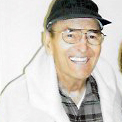 A man wearing a black baseball cap, glasses, a grey, plaid, collared button up, and a white thick jacket, smiling brightly for a picture with a plain white background.
