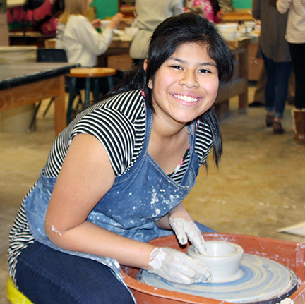 A young woman with black hair pulled back smiling brightly while wearing an apron and working at a pottery wheel making a bowl in a studio.