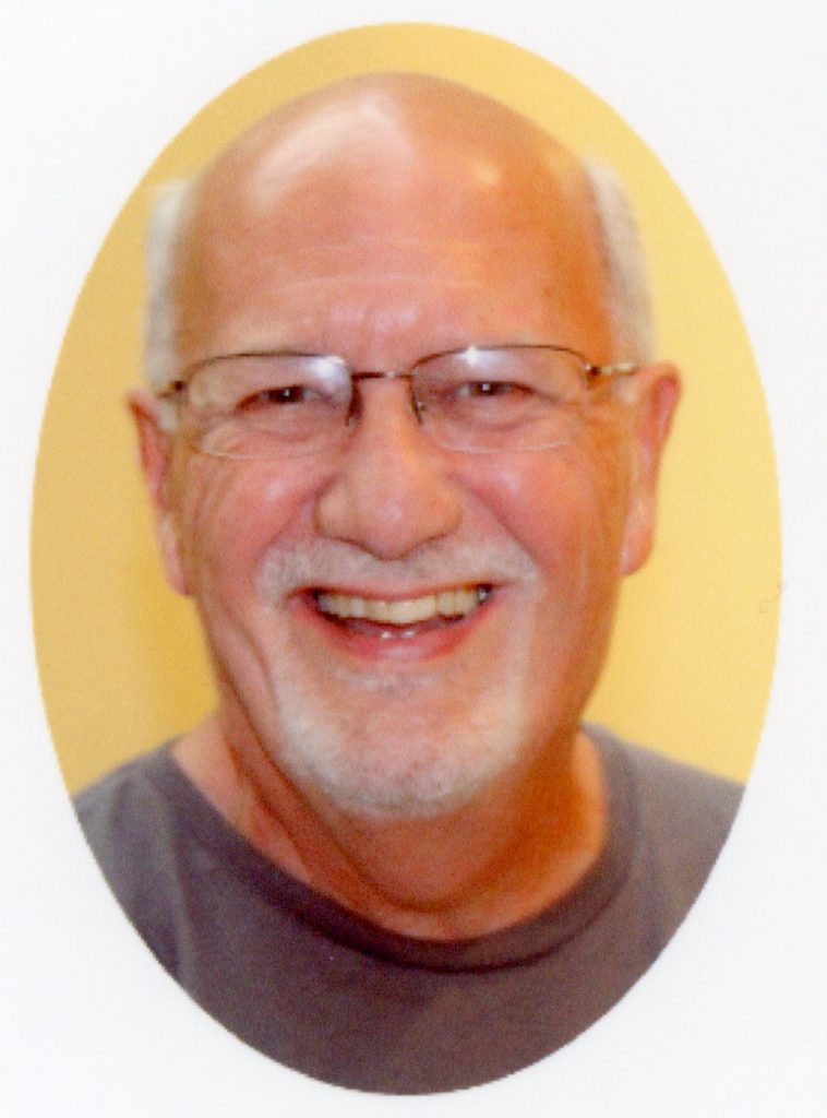 A man with grey hair and a grey beard smiling brightly for a portrait picture in a dark grey t-shirt and glasses with a light yellow background.