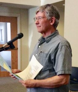 A man wearing glasses and a plaid button-up shirt standing indoors speaking into a microphone smiling with some papers in his hands.