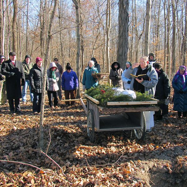 A priest blessing a corpse before it is lowered into it's grave in a wooded forest with numerous attendees surrounding the ceremony wearing big coats.