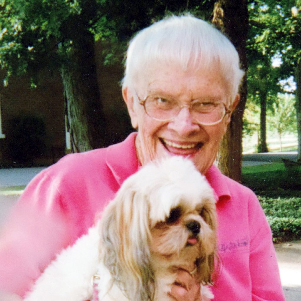 A woman with short white hair wearing a pink shirt smiling brightly for a picture outdoors while holding a small white dog with long grey ears.