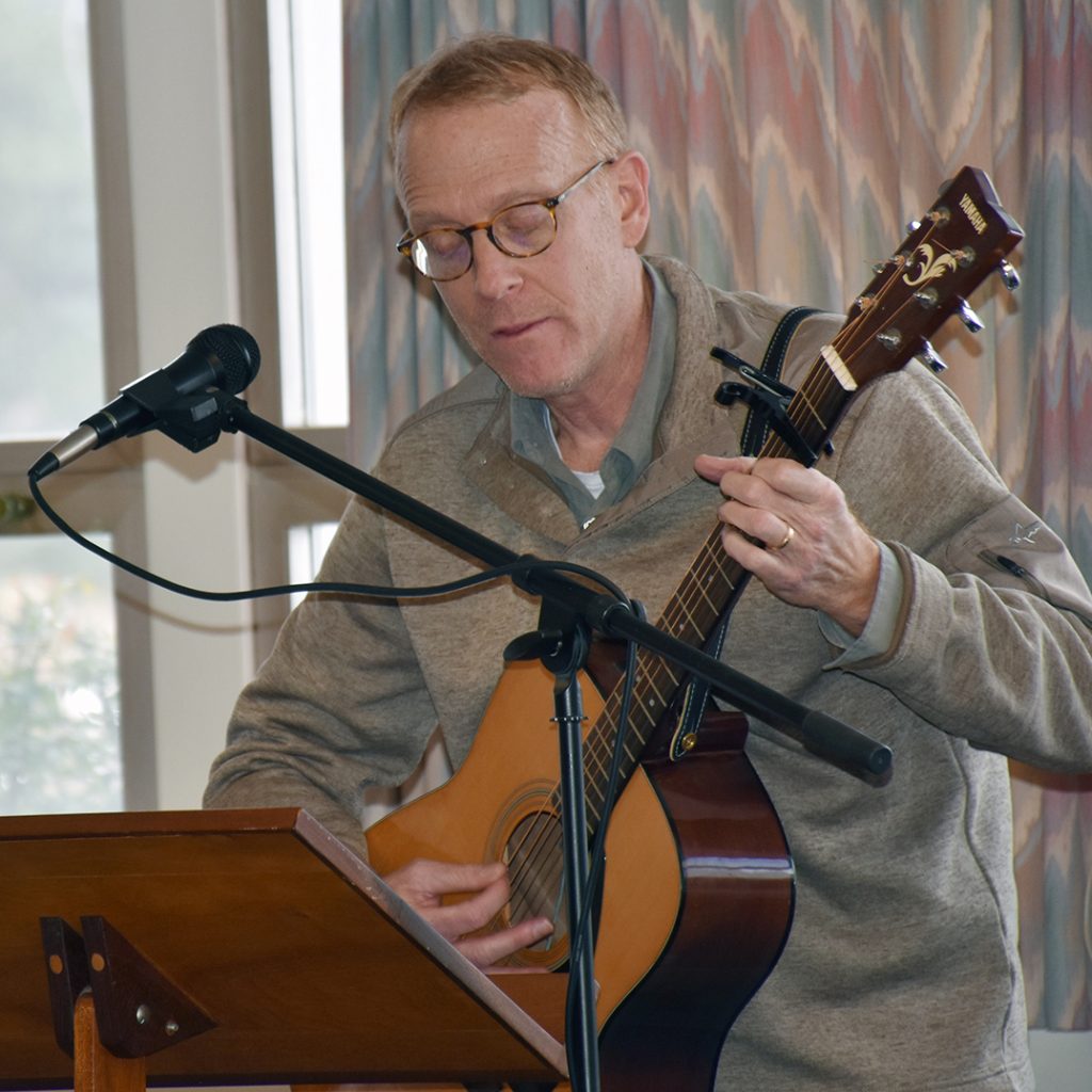 A man with glasses playing a guitar in front of a music stand and a microphone indoors.