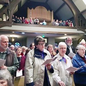 A choir sings in a two-story church chapel with a center organ. Many singers are wearing large coats and winter attire.