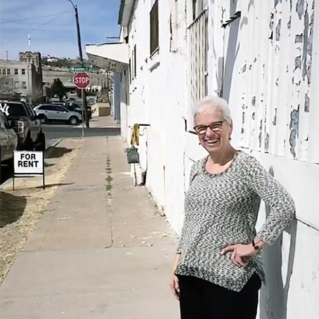 A woman with short grey hair and black-framed glasses smiling brightly underneath a white sign with black lettering that says: LORETTO on a white building outdoors on a sunny day.