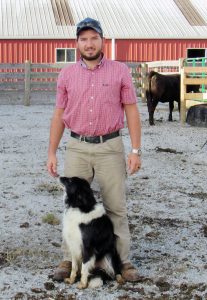 A man wearing a red flannel shirt, khakis, a baseball cap, and sunglasses, smiling with his black and white border collie on a farm in front of a red barn on a sunny day.