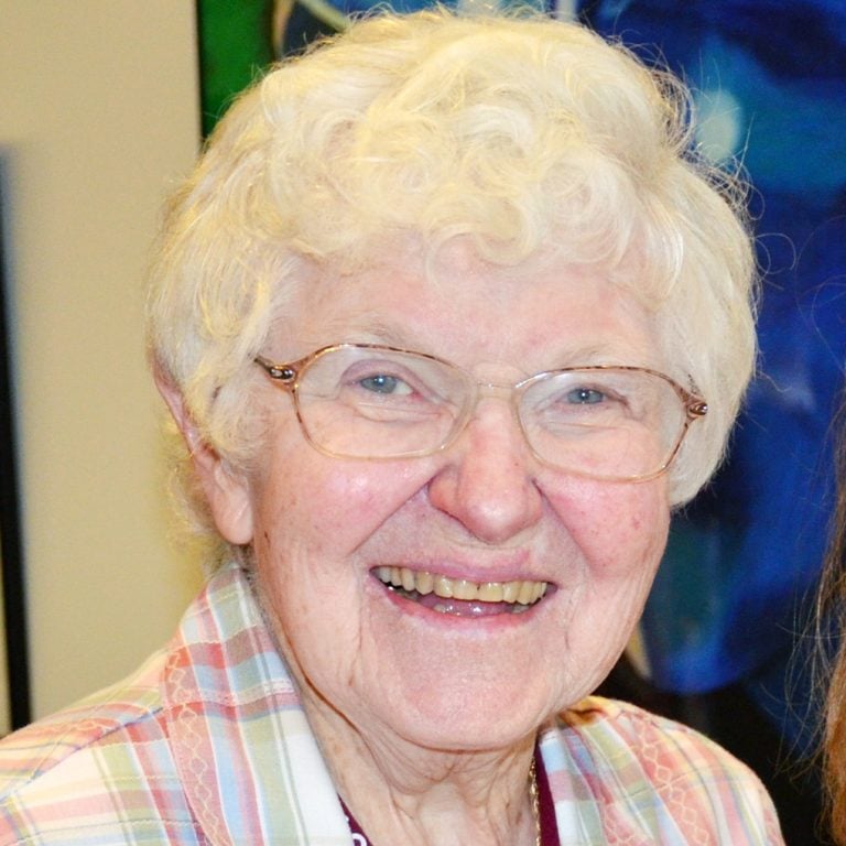 A woman with short, white hair, and oval glasses smiling brightly for a headshot picture indoors, wearing a light colored, plaid, collared, button up, and a red lanyard.