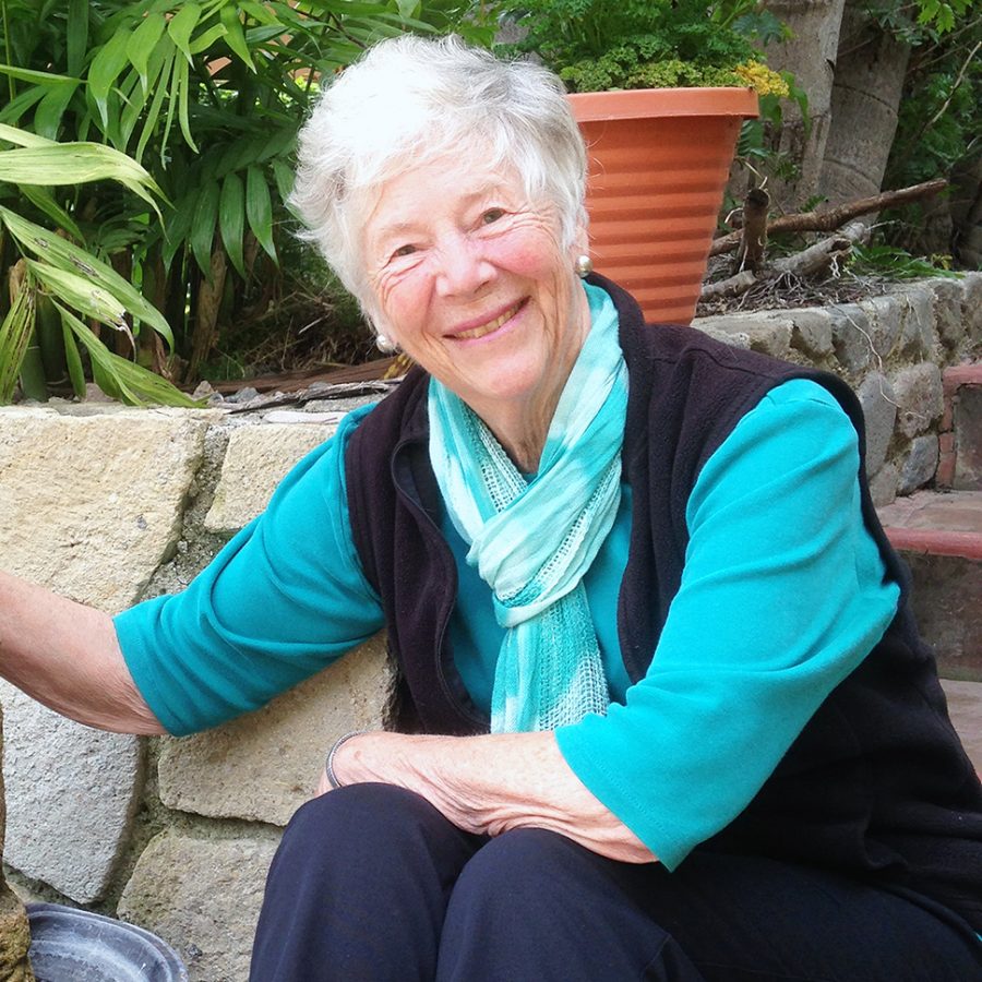 A woman with short grey hair smiling brightly for a portrait picture outdoors while she sits on stone stairs.