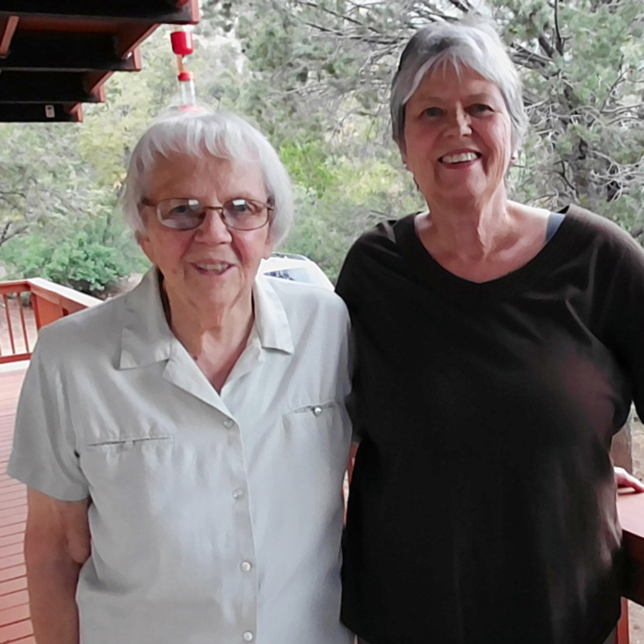Two women with short grey hair smiling brightly for a picture together on an outdoor balcony with big green trees behind them.