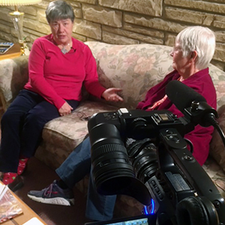 Two women sitting together and conversing on a couch in a living room while being interviewed on camera. The camera viewfinder is visible and one woman is looking directly into the camera.