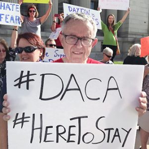 A man smiling brightly outdoors at a protest while holding a white sign with black text that says: #DACA #Here to Stay