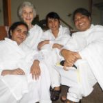 Four women wearing white robes smiling together for a group picture while sitting in a dark room on the floor.