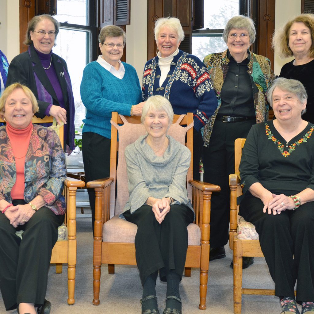 Four women sitting in chairs in a row with seven women standing behind them all smiling together for a group picture indoors.