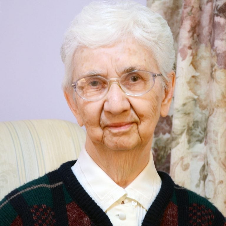 A woman with short, white hair and small, round glasses smiling for a headshot picture wearing a white collared blouse and a dark plaid sweater, sitting on a striped couch in a room.rose