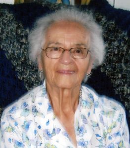 A woman with short grey hair wearing a white collared shirt with light blue and green patterns, and glasses smiling for a picture with a dark background.