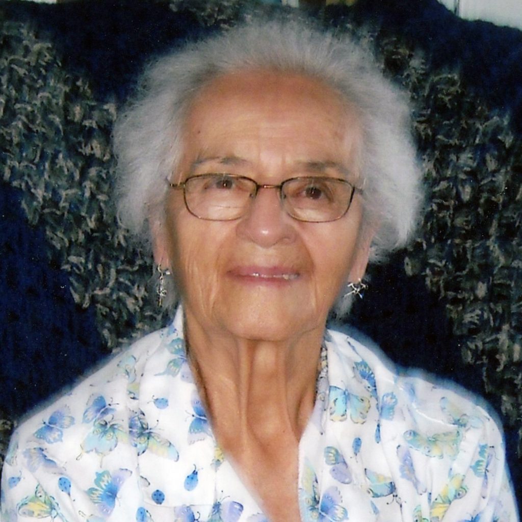 A woman with short grey hair wearing a white collared shirt with light blue and green patterns, and glasses smiling for a picture with a dark background.