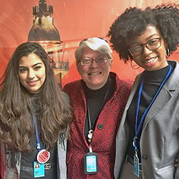 Four women smiling together while standing indoors. They are all wearing matching blue lanyards indicating they are at an event.