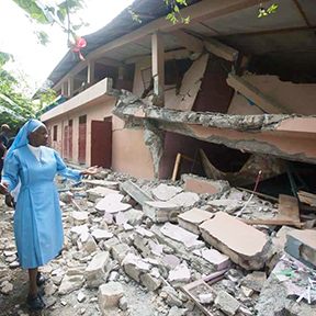 A nun wearing a blue dress and headdress displaying a destroyed building following an earthquake.