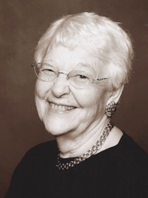 A black and white portrait picture of a woman smiling brightly with short white hair, and round wire glasses, wearing a chain necklace, earrings, and a black blouse with a dark grey background.