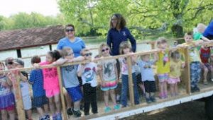 A group of young children lined up against a fence with two adult chaperones smiling together for a group picture outdoors on a sunny day.