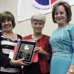 Three women smiling together for a group picture indoors while two of them hold a Hall of Fame plaque together.