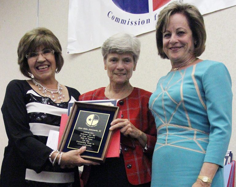 Three women smiling together for a group picture indoors while two of them hold a Hall of Fame plaque together.