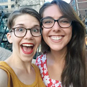 Two young women, both with dark-framed glasses, one with short brown hair and the other with long brown hair smiling together brightly for a selfie outdoors in a city.