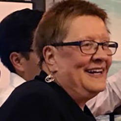 A woman with short brown hair and dark-framed glasses smiling candidly while looking to the side at a social event.