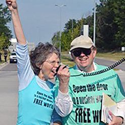 A woman speaking passionately on her friend's radio speaker outdoors. They are both wearing bright blue graphic t-shirts with bold black text.