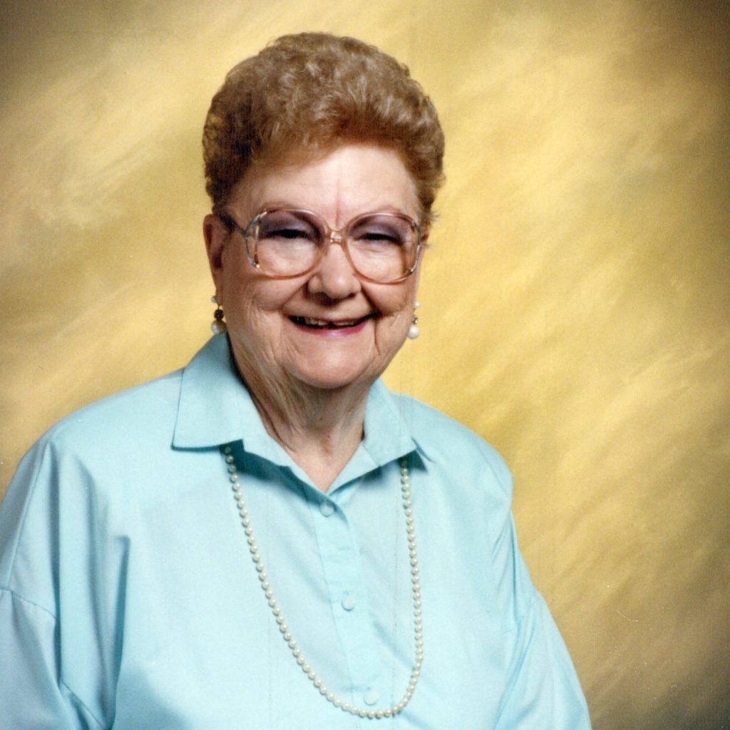 A portrait picture of a woman with short, light brown hair and glasses, wearing a baby blue collared button up and a long, gold necklace with a yellow background.