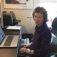 A woman with short brown hair smiling brightly for a picture while sitting at a desk with a laptop in an office.
