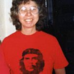 A woman with brown curly hair and big glasses smiling for a picture inside wearing a red shirt with a black graphic face.