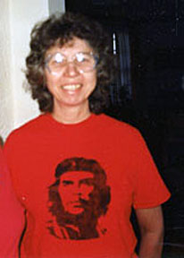 A woman with brown curly hair and big glasses smiling for a picture inside wearing a red shirt with a black graphic face.